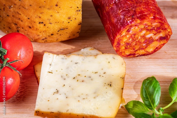 Fototapeta Sliced kulen and semi-hard cheese arranged neatly on a plate, garnished with fresh basil leaves. The vibrant colors of the red cured sausage, creamy-yellow cheese, and green basil create a visually ap