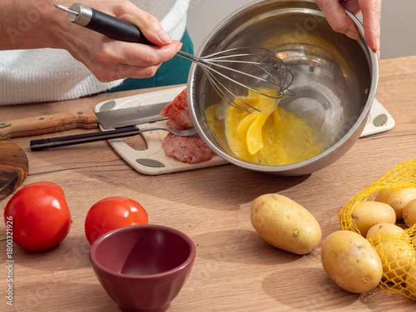 Obraz Close-up of a woman whisking eggs in a metal bowl while preparing a meal. Fresh vegetables and raw meat on the table. Authentic home cooking atmosphere.