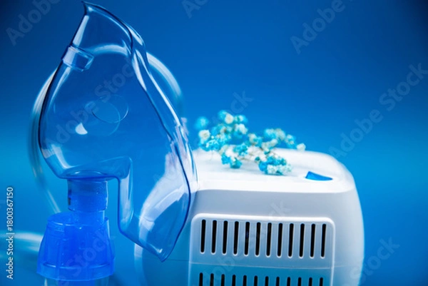 Fototapeta Close-up side view of a blue mask and a device on a blue background and flowers nearby. The concept of treating respiratory diseases, lung diseases, prevention and self-care. High quality photo