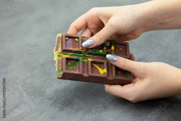 Obraz Female hands breaking off piece of Dubai chocolate on gray background. Green kadayif and pistachio spread with pistachio in trend dessert. Close-up, selective focus. High quality photo