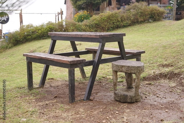 Obraz Rustic wooden picnic table with attached benches and a unique stone stool stands on a grassy patch, offering a tranquil outdoor seating area in a natural park