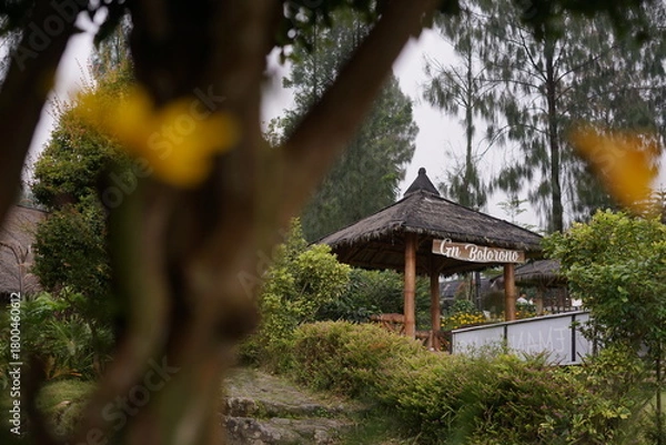 Obraz A tranquil rustic gazebo with a traditional thatched roof, nestled amidst lush green foliage and trees in a serene garden setting under an overcast sky, offering a peaceful natural retreat