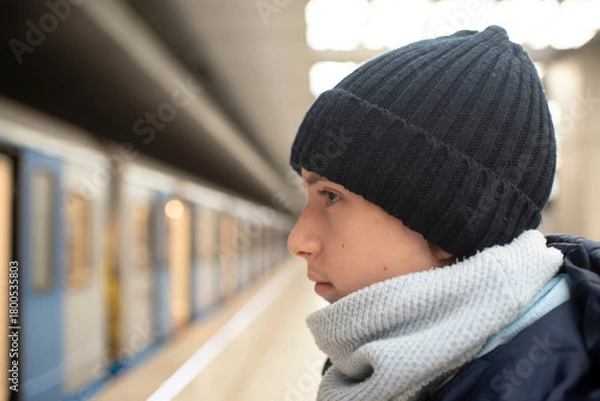 Fototapeta Profile of a teenage boy in a black hat, against the background of a train and a subway platform.