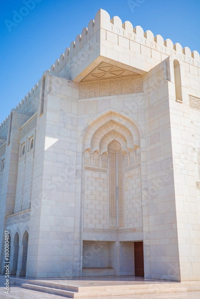 Fototapeta A bright architectural photograph capturing the white marble exterior of the Sultan Qaboos Grand Mosque in Muscat, Oman. The image features intricate Islamic carvings, an ornate arched doorway