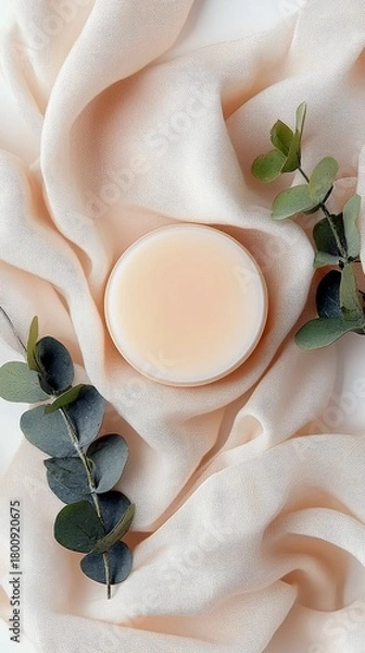 Fototapeta A cosmetic product, likely a balm or cream, is presented on a soft, flowing fabric with a neutral tone, accented by sprigs of eucalyptus.