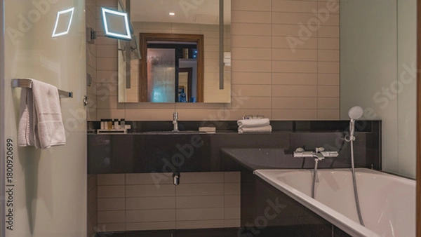 Obraz Bathroom interior. White rectangular bathtub, metal faucet, shower. The walls are tiled. Towels are on the counter in a roll, toiletries are laid out. Wall mirror.
