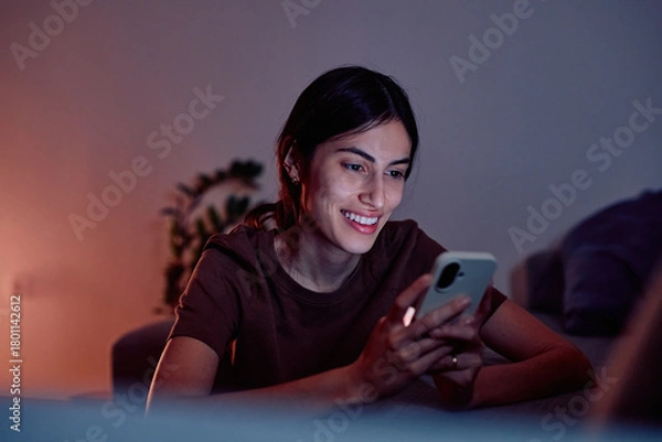 Fototapeta Young woman smiling while looking at her smartphone screen at home in low light, enjoying digital communication and apps