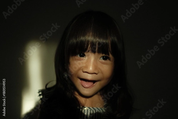 Obraz Portrait of a young girl standing in low-key lighting with a beam of natural light illuminating part of her face. She looks to the side with a calm expression, creating a cinematic