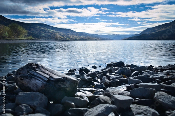 Obraz Clear mountain lake with rocky shore and distant view of mountains under blue sky
