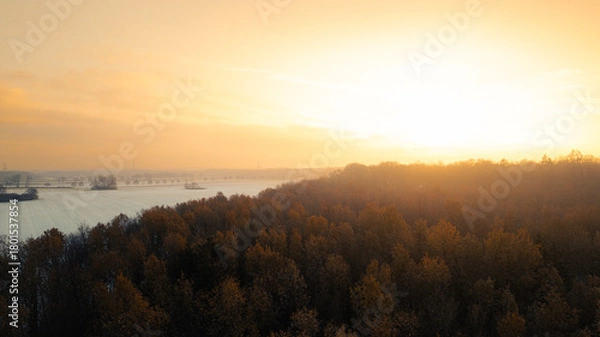 Fototapeta Atmospheric aerial view at sunrise with warm golden light illuminating an autumn forest and a river in the background. The morning fog and the glowing horizon create a calm, peaceful mood in this scen