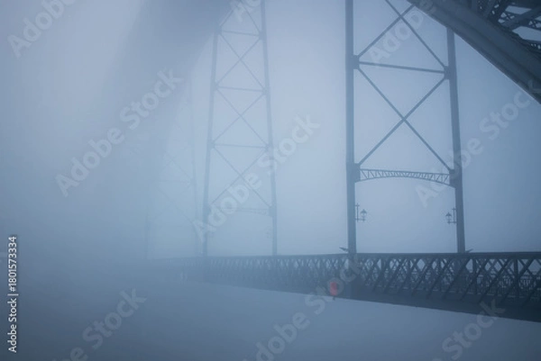 Obraz A dense fog envelops the Dom Luís I Bridge in Porto, Portugal, softening its steel structure into faint silhouettes. The scene feels quiet, surreal, and almost weightless.