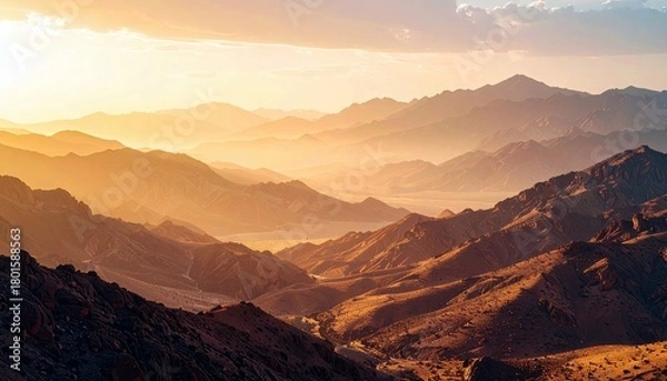 Fototapeta A vast expanse of layered mountain ranges is illuminated by the warm, golden light of sunset or sunrise, casting long shadows and creating a sense of depth and