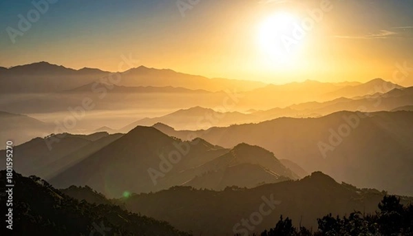 Fototapeta A breathtaking sunrise casts a warm golden glow over multiple layers of silhouetted mountain ranges, with mist filling the valleys below.