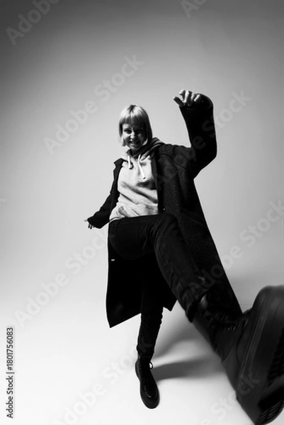 Fototapeta A person with light hair energetically kicks one leg forward, wearing a cozy hoodie and a long coat. The minimalist background highlights their joyful expression and dynamic movement