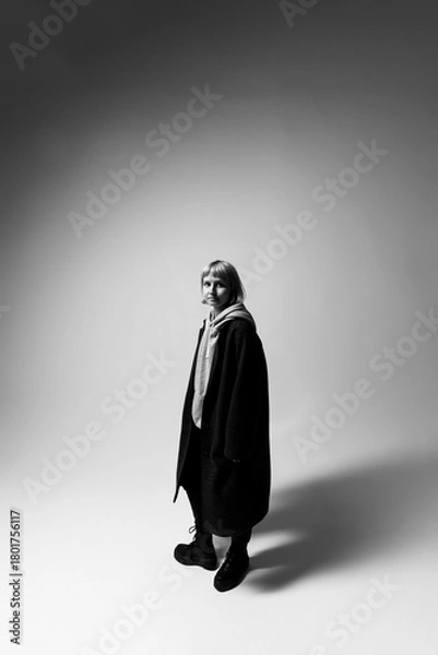 Fototapeta A woman stands in a stark, minimalist setting, dressed in a long coat and comfortable footwear. Her thoughtful gaze and relaxed stance convey a blend of confidence and ease