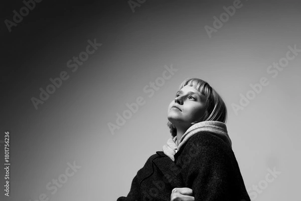 Fototapeta A young woman stands thoughtfully, her expression reflecting deep contemplation as she gazes upward against a soft, neutral background. The light creates a calm atmosphere