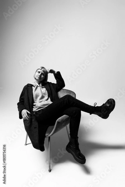Fototapeta A person sits confidently in a sleek chair, wearing a cozy hoodie and stylish coat. Their pose exudes a sense of relaxed sophistication against a neutral backdrop