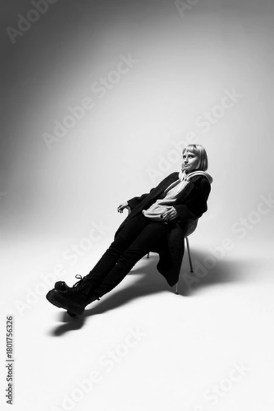 Fototapeta A fashionable young woman leans back casually on a chair, dressed in all black. The soft lighting creates a striking contrast, enhancing her relaxed yet confident pose