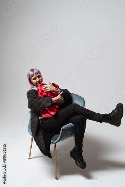 Fototapeta A cheerful woman sitting comfortably in a chic chair embraces a bright red blanket. She has short purple hair and wears a fashionable outfit, creating a lively atmosphere in a minimalist space
