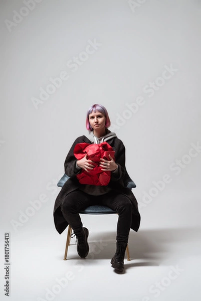 Fototapeta A person with pastel pink hair sits calmly on a stylish chair, cradling a bright red heart-shaped object. The background is simple and clean, focusing on the unique expression and pose