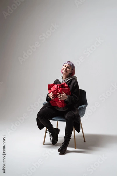 Fototapeta A person with purple hair smiles brightly while seated in a stylish chair. They hold a large red gift close, expressing excitement and joy in a simple, minimalist setting