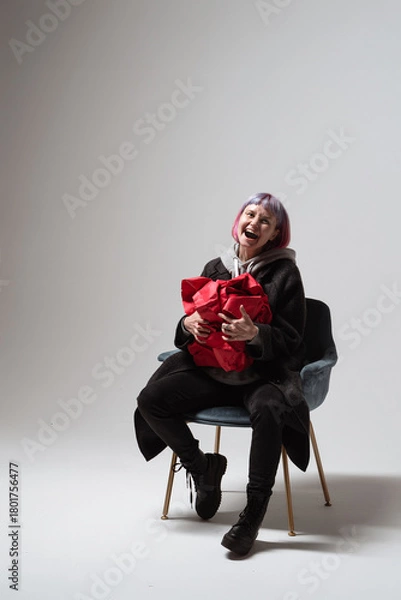 Fototapeta A woman with short pink hair sits on a stylish chair, joyfully holding a red gift close to her chest. Her laughter fills the minimalist space, creating a warm and cheerful atmosphere