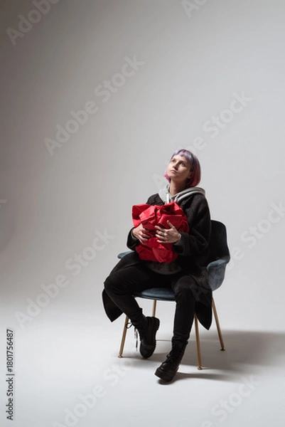 Fototapeta In a minimalist studio, a young person sits in a stylish chair, cradling a vibrant red object. Their expression reflects a deep contemplation, illuminated by gentle light