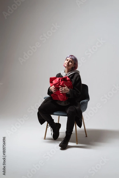 Obraz A woman wearing a casual outfit sits comfortably in a chair, cradling a vibrant red gift. Her expression radiates happiness as she enjoys this delightful moment of anticipation