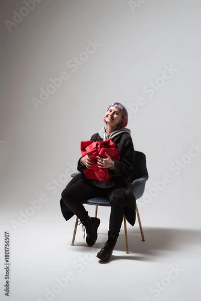 Fototapeta A person with pastel hair smiles brightly while holding a striking red gift close. Seated on a modern chair in a simple setting, the scene captures pure happiness and excitement