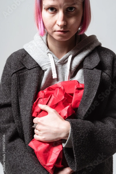 Fototapeta A person stands against a simple background, holding a crumpled red paper tightly. Their expression is serious, and their pink hair pops against the gray coat they wear