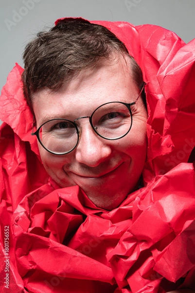 Fototapeta In a colorful studio, a young man joyfully poses with his face wrapped in vibrant red paper. His playful expression captures the essence of fun and creativity in this lively moment