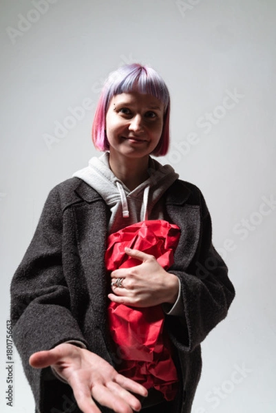 Fototapeta In a minimalistic studio, a smiling person with colorful hair holds a wrapped gift close to their chest. Their open hand suggests a welcoming gesture, enhancing the cheerful atmosphere