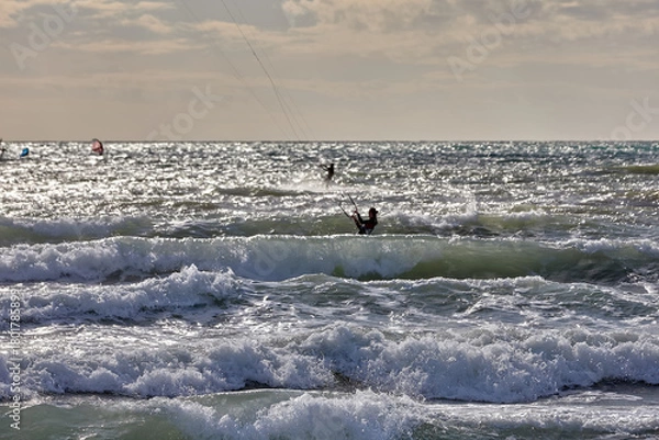 Fototapeta Kitesurfer glides through strong waves under an overcast sky, surrounded by the shimmer of sunlight on the restless sea. motion, energy