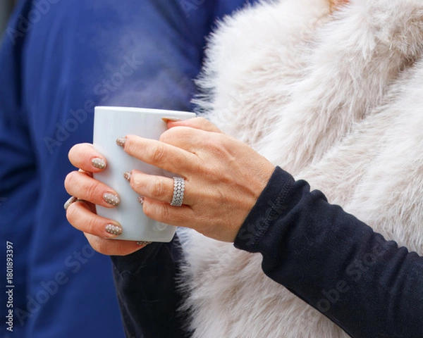 Fototapeta A visitor at the autumn Naplavka market holds a warm drink in a white mug, hands adorned with glittery nails and rings, capturing the cozy atmosphere and seasonal charm of this popular riverside event