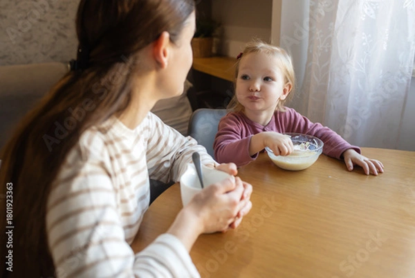 Fototapeta Happy single mother doing her daughter's hair while having breakfast. Woman combs the hair of a little girl. The concept of morning routine, single parenthood and preparation for school