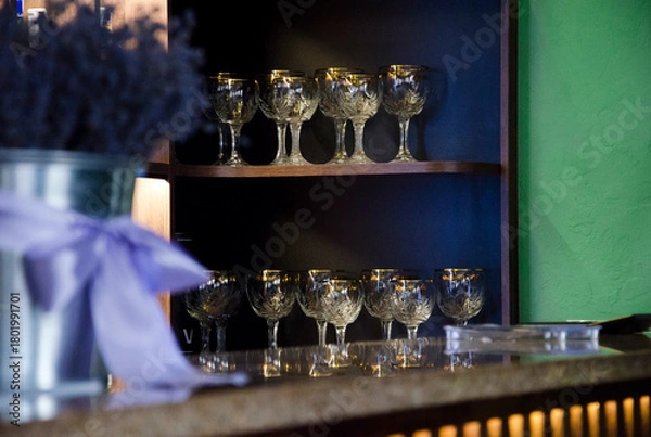 Fototapeta Collection of elegant crystal glasses with gold rims is neatly arranged on wooden shelves in a stylish bar interior.