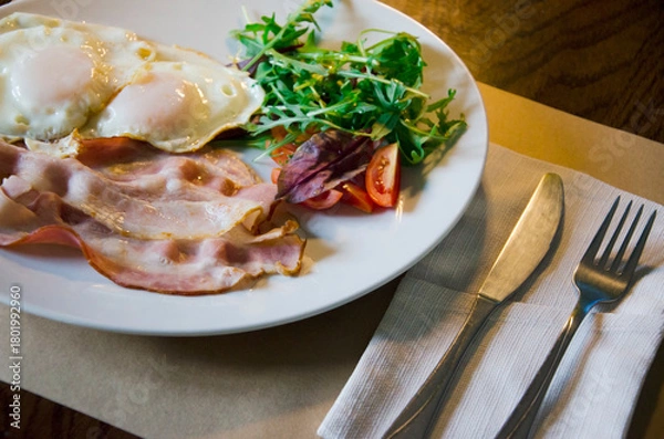Fototapeta Classic breakfast plate featuring fried eggs, crispy bacon, fresh greens, and sliced tomatoes served on a white dish.