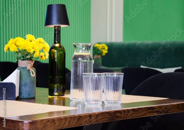 Fototapeta A stylish café table set with a glass water carafe, two drinking glasses, and a decorative bottle lamp.