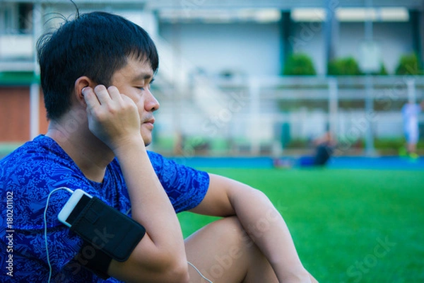 Fototapeta Asian sport man is listening his favorite song by smart pohne that hold on his arm during relax in sport ground.
