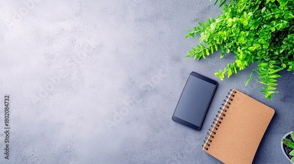 Obraz A modern workspace setup featuring a smartphone, a spiral-bound notebook, and a lush green plant on a textured gray background.