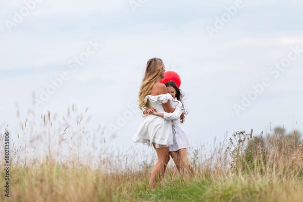 Fototapeta Mother and child enjoying a playful moment outdoors with a red balloon in a grassy field on a cloudy day