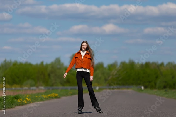 Fototapeta Young woman skating confidently on an empty road surrounded by green trees under a blue sky