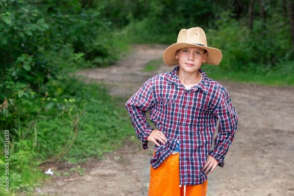 Obraz Young boy in a straw hat stands proudly on a forest path surrounded by greenery during daytime