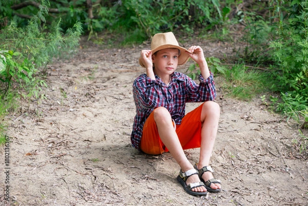 Obraz A young boy sits on the ground in a forest, dressed in a plaid shirt and bright orange shorts. He wears a straw hat and looks contemplative, surrounded by nature.