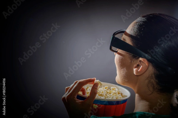 Fototapeta A woman wearing 3D glasses is viewed from the side, illuminated by the light of a screen as she eats popcorn from a striped bucket in a dark theater or home cinema setting.