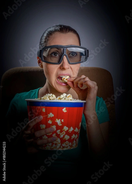 Fototapeta A woman wearing 3D glasses sits in a dark room, intently watching a screen while vigorously eating popcorn from a large red and blue bucket.