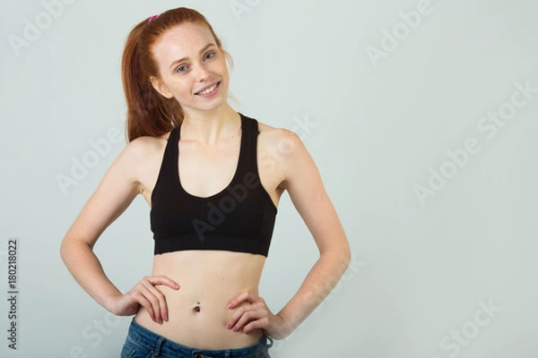 Fototapeta beautiful slender girl with a smile with red hair in sportswear on a light background
