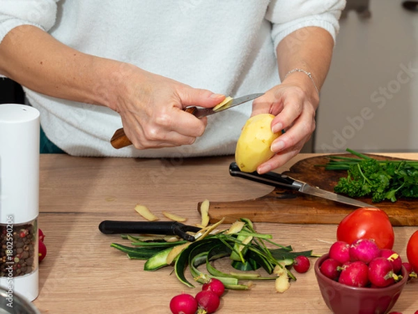 Obraz Woman peeling a potato with a knife on a kitchen counter, surrounded by fresh vegetables and herbs. Home meal preparation.