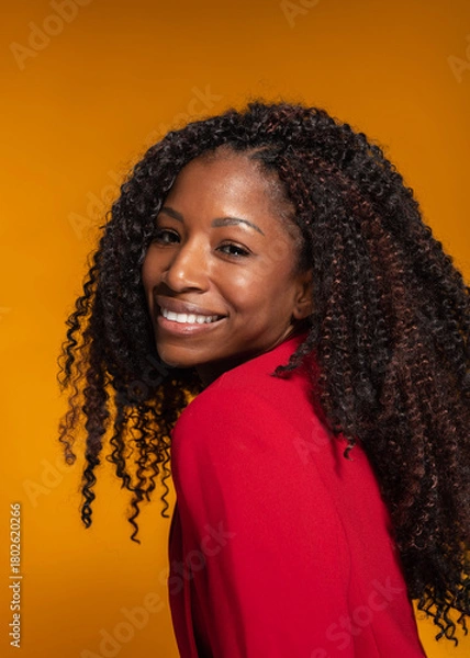 Obraz Studio shot of cheerful Hispanic woman in red blazer smiling over her shoulder on yellow background