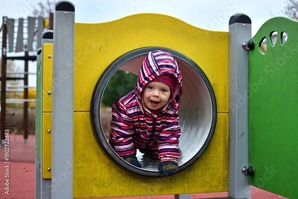 Fototapeta Smiling toddler in a colorful striped snowsuit playing inside a playground tunnel on an autumn day. Bright yellow structure and soft light capture a joyful, candid childhood moment.